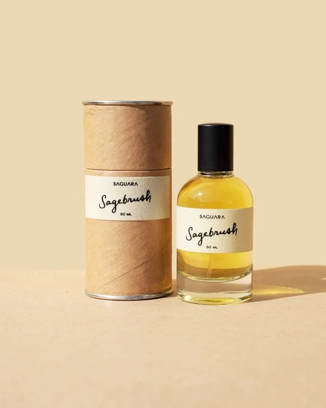 A clear glass bottle labeled "Sagebrush | Saguara Perfumes" (50 mL Eau de Parfum, vegan, with California sage notes) and its cylindrical cardboard container from Saguara Perfumes are displayed on a beige surface and background.