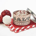 An open tin of Santa Sea Salt by Beautiful Briny Sea, filled with a festive blend of organic rosemary and pink peppercorn, sits on red and white napkins beside three decorative holiday balls.