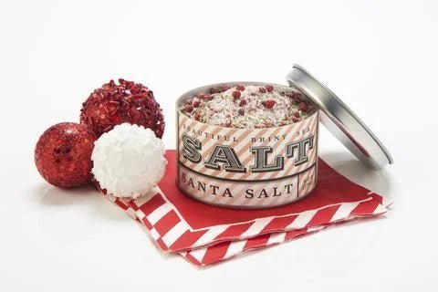 An open tin of Santa Sea Salt by Beautiful Briny Sea, filled with a festive blend of organic rosemary and pink peppercorn, sits on red and white napkins beside three decorative holiday balls.