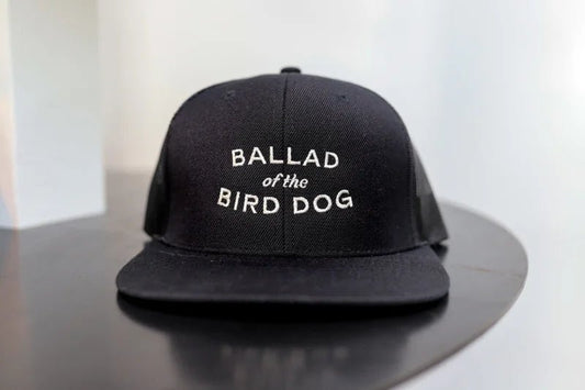 The Shop Hat from Ballad of the Bird Dog features a black snapback design with a white embroidered simple oval logo—ideal for fans of classic branding and stylish caps.
