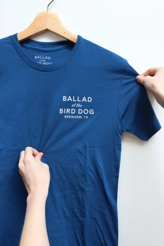 Person Holding T-shirt With ’bald’ Word Displayed In Shop Shirt | Bird Dog Classic Shop Logo