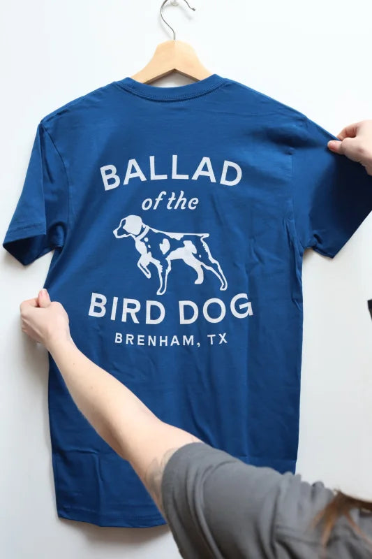 Woman Holding a Blue Shirt With ’ballad Of The Bird Dog’ Displayed In Shop Shirt Classic Logo.