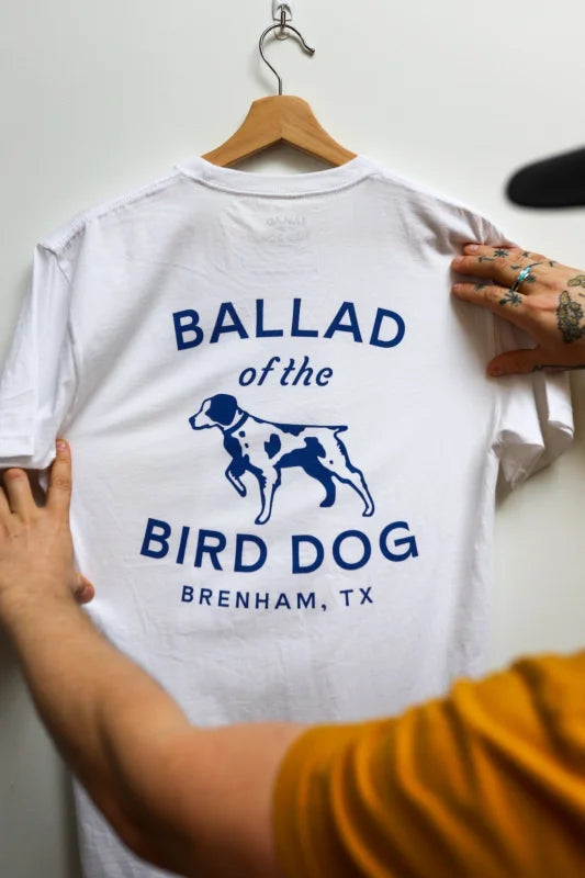 Man Holding White ’shop Shirt’ With Blue Lettering From Bird Dog Classic Shop Logo.
