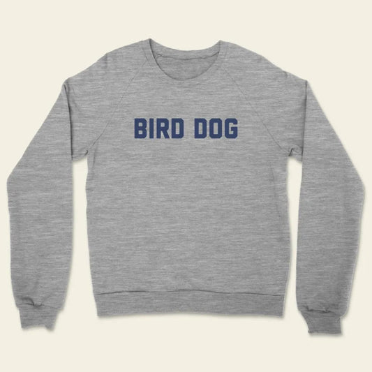 The Bird Dog Sweater by Ballad of the Bird Dog is a unisex crewneck, made in the USA, with "BIRD DOG" boldly printed in dark blue across soft gray fabric.