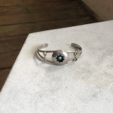 The Vintage Small Shadow Box Squash Blossom & Cuff, a sterling silver bracelet with a round dark star-shaped centerpiece, is displayed on a light-colored surface.