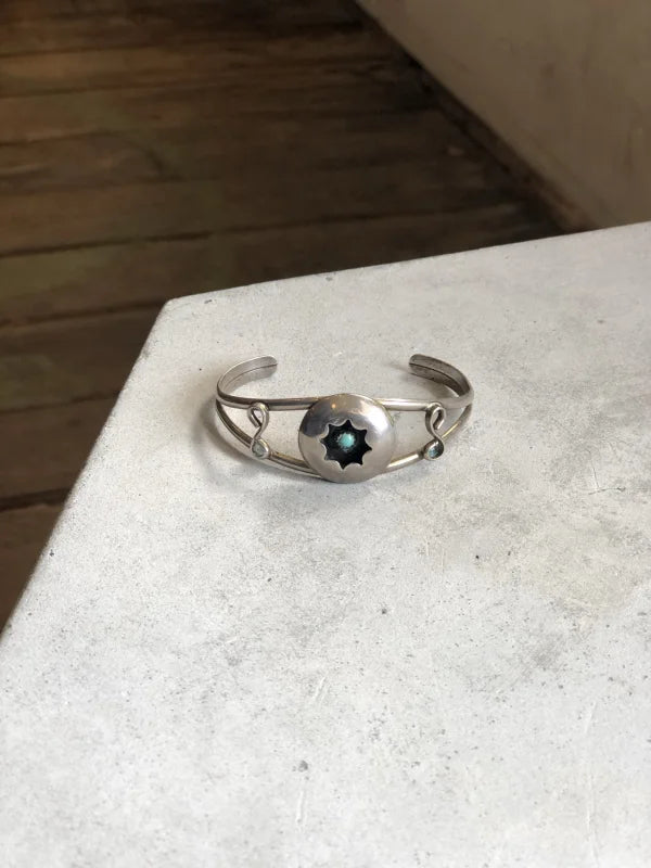 The Vintage Small Shadow Box Squash Blossom & Cuff, a sterling silver bracelet with a round dark star-shaped centerpiece, is displayed on a light-colored surface.