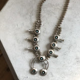 The Vintage Small Shadow Box Squash Blossom & Cuff, featuring turquoise stones and a subtle shadow box design, is displayed on a light-colored surface.