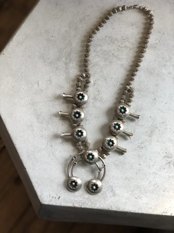 The Vintage Small Shadow Box Squash Blossom & Cuff, featuring turquoise stones and a subtle shadow box design, is displayed on a light-colored surface.