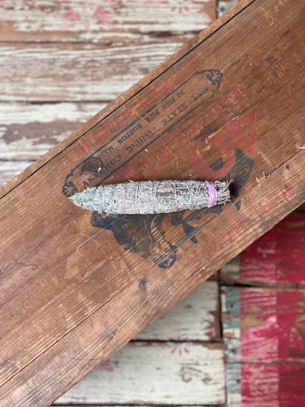 A Taos Herb Co. Smudge Stick | Seven Direction, wrapped with string, sits atop a weathered wooden surface marked by faded red and black lettering.