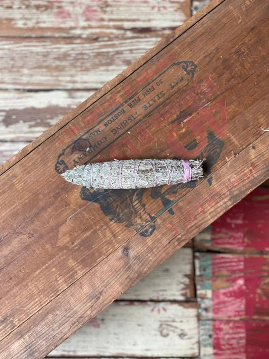 The Smudge Stick | Seven Direction by Taos Herb Co., wrapped with string, rests on a weathered wooden surface with faded red lettering, evoking the tradition of Native American blessings.