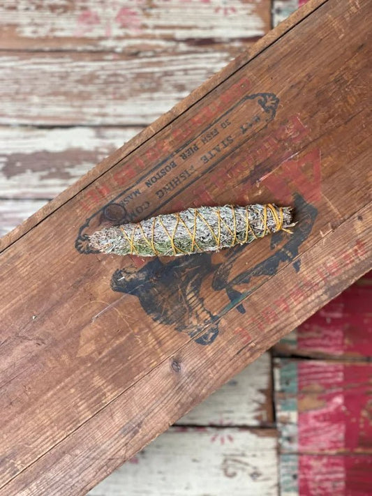 The Smudge Stick | Spirit Calling by Taos Herb Co., a cherished sacred herb wrapped in yellow string, rests atop a vintage wooden cigar box with faded graphics.