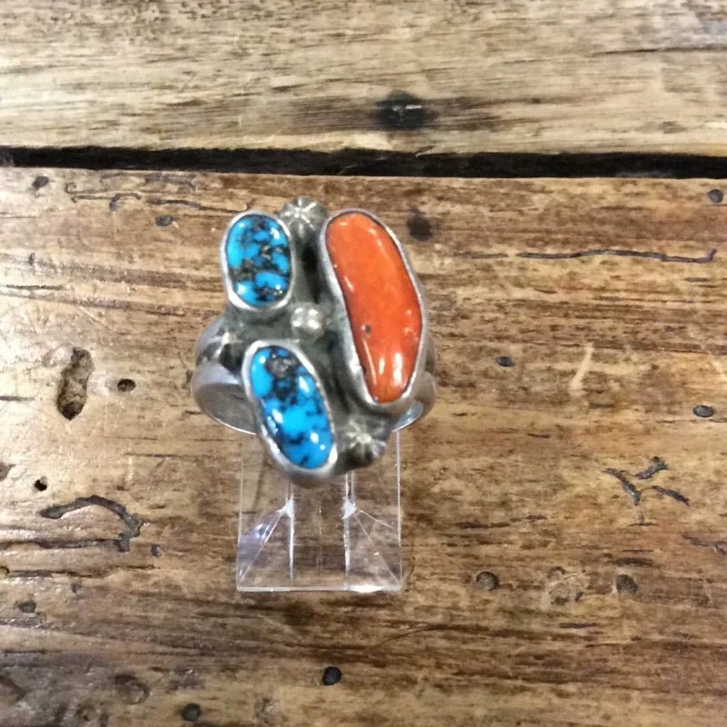 The Vintage Three-stone Turquoise & Coral Ring, featuring two turquoise stones and a vibrant coral stone, is displayed on a clear stand atop a rustic wooden surface.