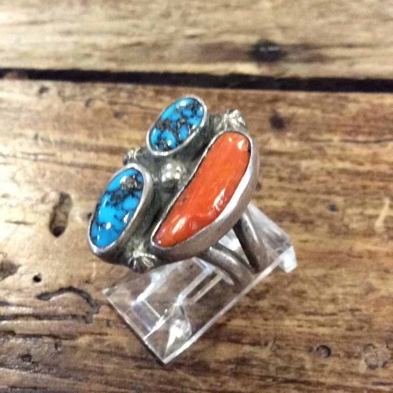 The Vintage Three-stone Turquoise & Coral Ring features two oval turquoise stones and an elongated orange coral stone, presented on a clear stand over a wooden surface.