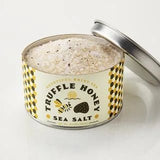 An open tin of Beautiful Briny Sea's Truffle Honey Sea Salt is filled with coarse, speckled salt infused with Italian black truffles, and its lid sits beside the container.