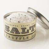 A metal tin of Truffle Sea Salt by Beautiful Briny Sea, filled with coarse white salt and Italian black truffle pieces, sits on a white surface with its lid resting behind it.