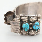 The Turquoise Watch Cuff by Vintage is a sterling silver cuff adorned with two turquoise stones set amid detailed leaf and feather designs.