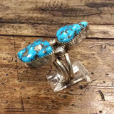 The Vintage Two-stone Turquoise Ring, size 8.5, features two large irregular turquoise stones set in sterling silver and is displayed on a clear stand atop a rustic wooden surface.