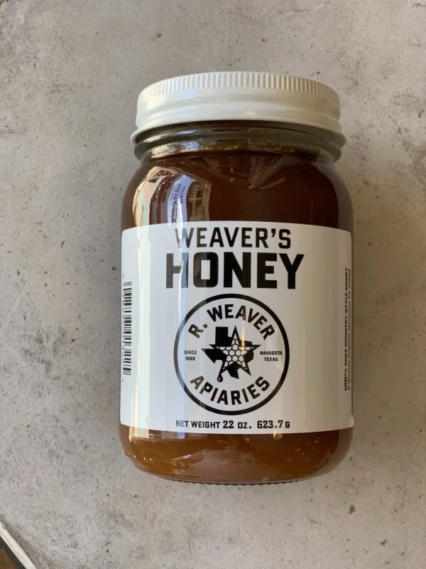 A 22-ounce jar of Weaver's Honey by R. Weaver Apiaries, featuring a white label with the R. Weaver Apiaries logo and text, sits on a light surface—a true taste of Texas honey.