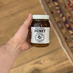 A hand holds a jar of Weaver's Honey by R. Weaver Apiaries, featuring a white label and lid, over a wooden floor next to a patterned rug.