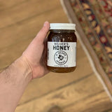 A hand holds a jar of Weaver's Honey by R. Weaver Apiaries, featuring a white label and lid, over a wooden floor next to a patterned rug.