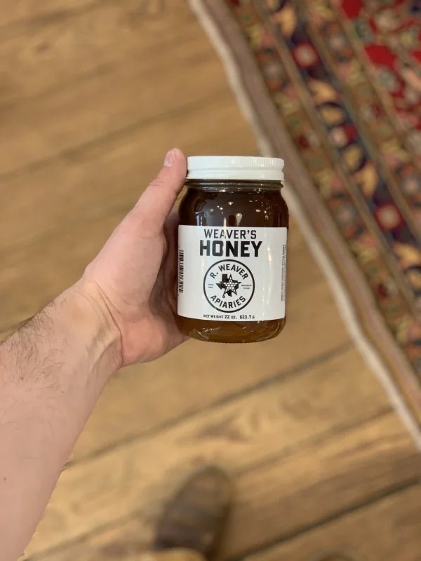 A hand holds a jar of Weaver's Honey by R. Weaver Apiaries, featuring a white label and lid, over a wooden floor next to a patterned rug.