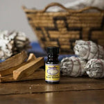 A small bottle of essential oil stands on a wooden surface next to Taos Herb Co. White Sage Smudge Stick, palo santo, and a woven basket in the background.