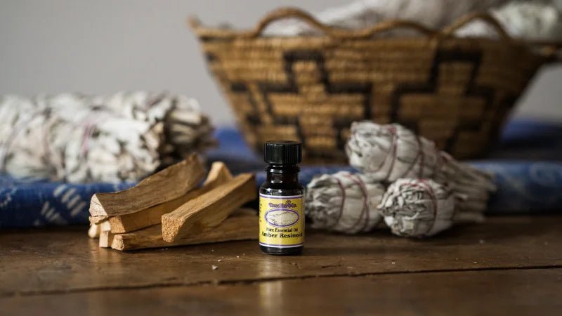 A small bottle of essential oil stands on a wooden surface next to Taos Herb Co. White Sage Smudge Stick, palo santo, and a woven basket in the background.