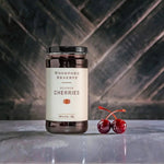 A jar of Woodford Reserve Bourbon Cherries by Bourbon Barrel Foods rests on marble beside two fresh cherries—an ideal garnish for your next Manhattan—against a dark, patterned backdrop.