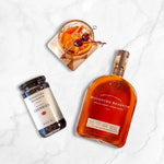 A bottle of Woodford Reserve bourbon, a jar of Bourbon Barrel Foods' Woodford Reserve Bourbon Cherries, and a Manhattan cocktail garnished with these cherries are arranged on a white marble surface.