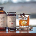 A bottle of cocktail syrup, a jar of Woodford Reserve Bourbon Cherries by Bourbon Barrel Foods, and a glass of Manhattan Cocktail garnished with cherries are arranged on a table.