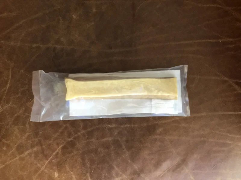 A sealed pouch from Rogue Pet Feed contains a rectangular yellowish Yak Stick high-protein dog treat, placed on folded white paper and set on a brown leather surface.