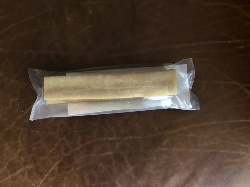 The Yak Stick by Rogue Pet Feed is a sealed, cylindrical dog treat made of light-colored material in plastic packaging, shown on a brown leather surface—a high-protein snack for your pup.