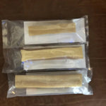 Three vacuum-sealed packages of Rogue Pet Feed Yak Stick dog chews, each containing a single tan, cylindrical stick, are arranged in a vertical row on a brown surface.