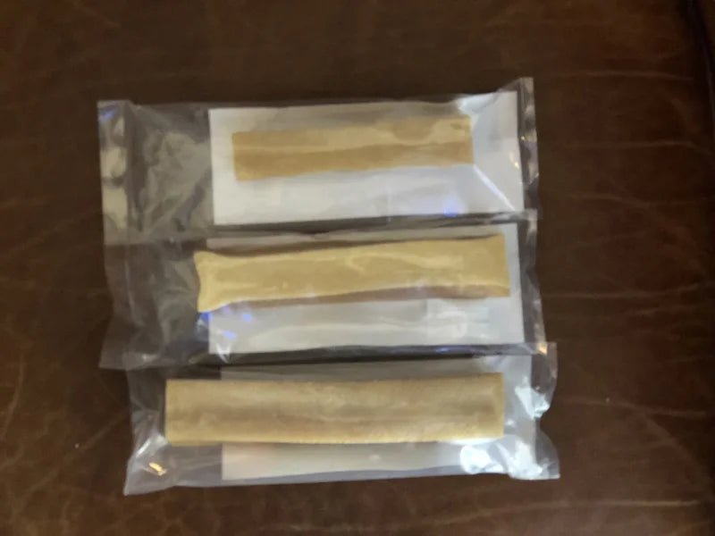 Three vacuum-sealed packages of Rogue Pet Feed Yak Stick dog chews, each containing a single tan, cylindrical stick, are arranged in a vertical row on a brown surface.