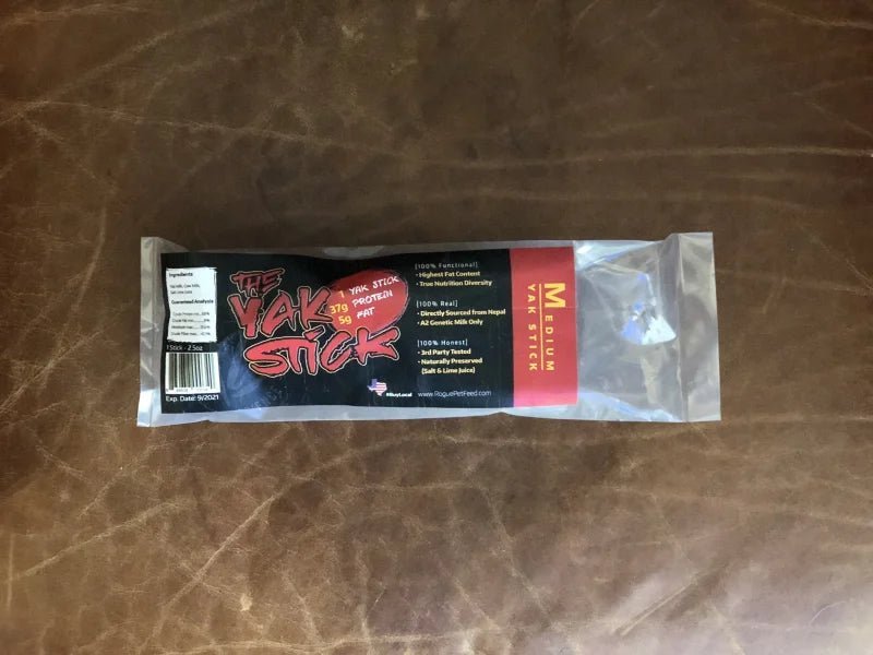 A packaged dog chew labeled "Yak Stick | Rogue Pet Feed" from Rogue Pet Feed sits on a brown leather surface, with packaging highlighting its high protein content and product details.