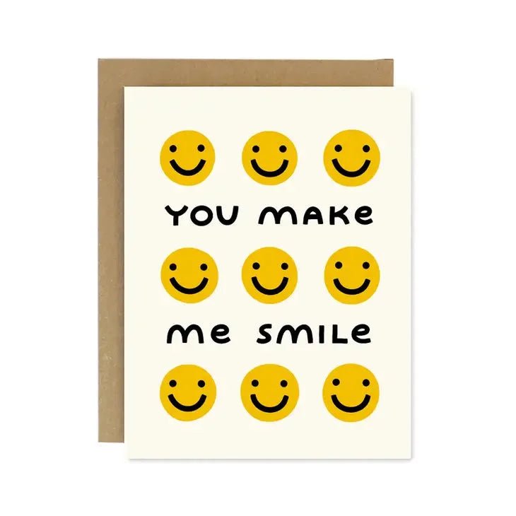 The Worthwhile Paper "You Make Me Smile" card features nine yellow smiley faces and bold central text. Made from recycled paper, this greeting card comes with a brown envelope partially visible behind it.